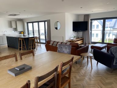 An open plan kitchen and living room with a wooden dining table and chairs a kitchen island with stools leather sofas a wall mounted TV and large windows at Galwad y Mor in Broad Haven