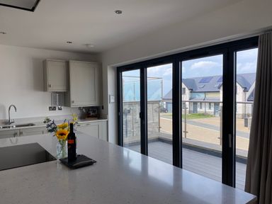A kitchen with a countertop with flowers and a bottle next to sliding glass doors showing a balcony and houses outside at Galwad y Mor in Broad Haven