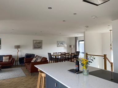 An open-plan living room and dining area with brown and gray chairs a dining table and a kitchen island with flowers and a bottle on top at Galwad y Mor Broad Haven