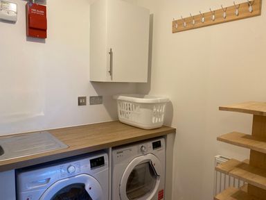 A laundry room with a washing machine and dryer under a wood countertop with a white laundry basket and hooks on the wall at Galwad y Mor in Broad Haven