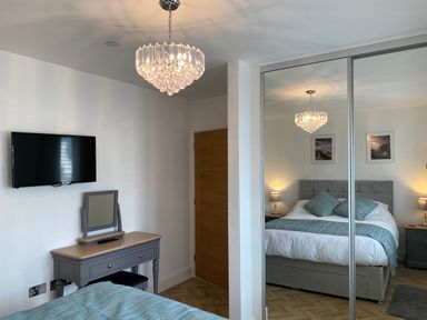 A bedroom with a bed and pillows bedside tables lamps a wall-mounted TV and a dresser with a mirror at Galwad y Mor in Broad Haven