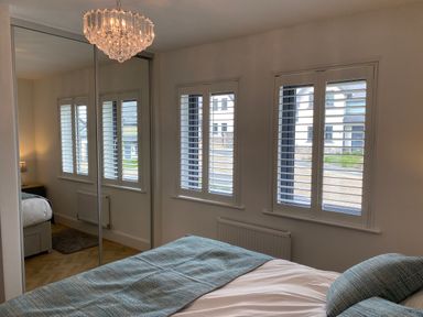 A bedroom with a bed covered with blue blankets white shutters on windows a mirrored wardrobe and a chandelier at Galwad y Mor in Broad Haven