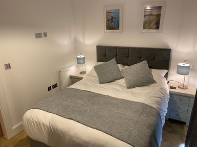 A bedroom with a double bed grey pillows and blanket two bedside tables with lamps and framed pictures on walls at Galwad y Mor in Broad Haven