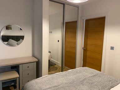 A bedroom with a bed covered by a gray blanket a small vanity with a stool and a round mirror two wooden doors and a mirrored sliding closet door at Galwad y Mor in Broad Haven