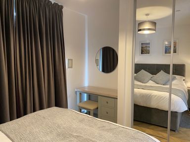 A bedroom with a bed covered with a gray blanket two pillows a dressing table with a circular mirror and a chair next to dark curtains at Galwad y Mor in Broad Haven