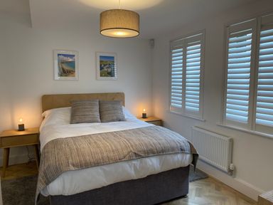A bedroom with a bed with pillows and a blanket two bedside tables with lamps three windows with shutters and two framed pictures on the wall at Galwad y Mor in Broad Haven