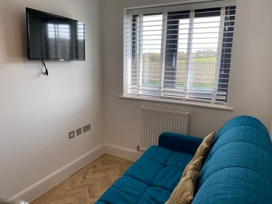 A small room with a blue sofa and two cushions a wall-mounted TV and a window with blinds at Galwad y Mor in Broad Haven