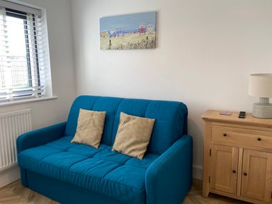 A blue sofa with two patterned cushions next to a wooden cabinet with a lamp and remote controls near a window with blinds at Galwad y Mor in Broad Haven