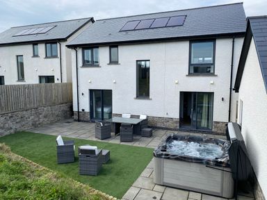 A patio area with outdoor furniture and a hot tub outside a modern white house with solar panels at Galwad y Mor in Broad Haven