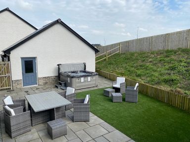 An outdoor patio area with gray wicker furniture including a table and chairs a hot tub on a paved surface and a grassy slope enclosed by a wooden fence at Galwad y Mor in Broad Haven
