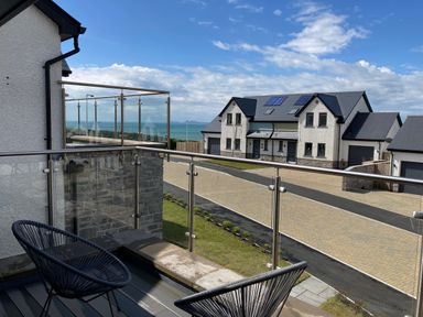 A balcony with two black chairs and glass railing overlooking a street with modern houses and a view of the sea at Galwad y Mor in Broad Haven