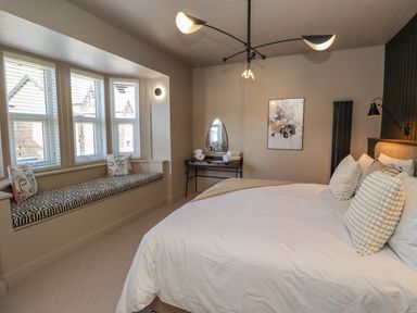 A bedroom with a large bed three pillows a window seat with cushions a vanity table with a mirror and rolled towels and floral artwork at Glencoe House in Wooler