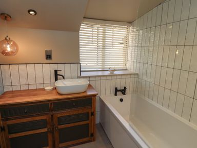 A bathroom with a wooden vanity with a white sink and black faucet next to a bathtub with black fixtures under a window with blinds at Glencoe House in Wooler