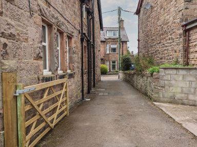 A narrow alleyway between stone buildings with a wooden gate and a house in the background at Glencoe House in Wooler