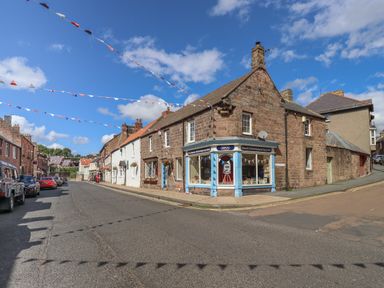 A street with parked cars decorated with hanging bunting and stone buildings including a corner shop at Glencoe House in Wooler