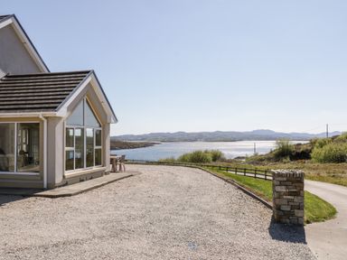 The exterior of a house with large windows next to a gravel driveway and a view of water and hills at Inverbeg Cottage 1 in Downings