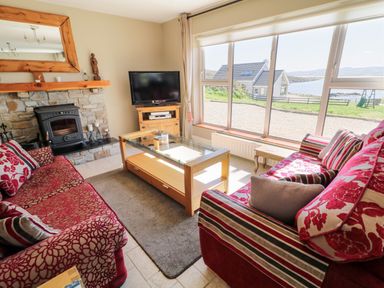 A living room with red patterned sofas a glass coffee table a stone fireplace and large windows overlooking a coastal view at Inverbeg Cottage 1 in Downings
