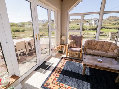 A sunlit room with wicker furniture a wooden coffee table and a patterned rug with glass doors opening to an outdoor dining area at Inverbeg Cottage 1 in Downings