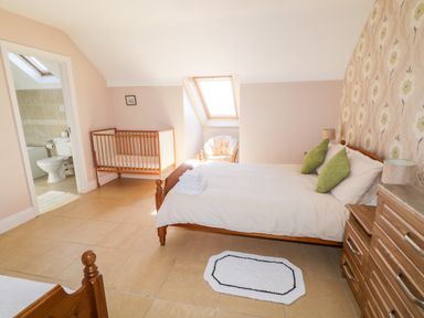 A bedroom with a double bed with green pillows a wooden crib a chair a wooden dresser and an open door to a bathroom at Inverbeg Cottage 1 in Downings