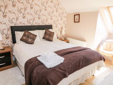 A bedroom with a bed with white sheets and brown pillows a brown throw and folded towels two bedside tables with lamps a window and a wicker chair at Inverbeg Cottage 1 in Downings