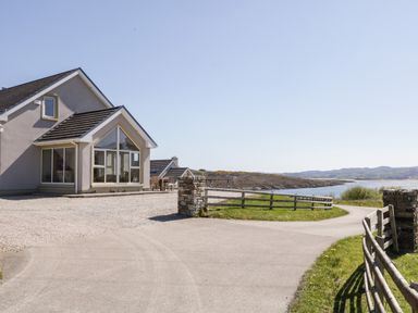 A house with large windows by a driveway and a wooden fence near a body of water at Inverbeg Cottage 1 in Downings