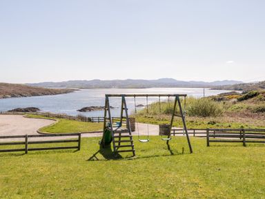A wooden swing set with two swings and a slide on grass near a body of water at Inverbeg Cottage 1 in Downings
