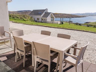 An outdoor patio with a table and six chairs overlooking a house a playground and a body of water at Inverbeg Cottage 1 in Downings