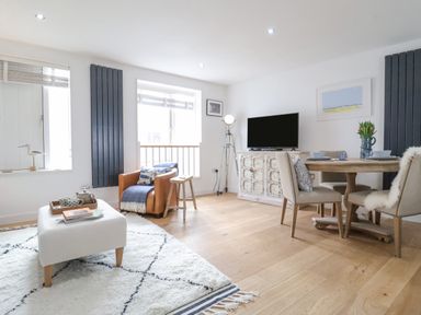 A living room with a dining table chairs a television on a cabinet an armchair and a white rug at The Gallivant in Southwold