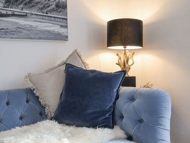 A blue tufted sofa with a blue and beige pillow a table lamp with a black shade and a wall picture at The Gallivant in Southwold
