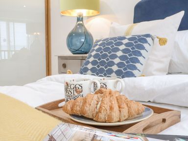 A bed with a wooden tray holding a plate with a croissant and two mugs near a blue patterned pillow and a lamp on a bedside table at The Gallivant in Southwold