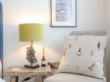 A bedside table with a lamp and diffuser next to a chair with a cushion and a framed picture on the wall at The Gallivant in Southwold