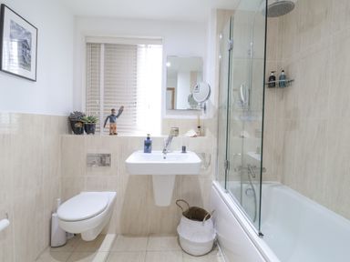 A bathroom with a wall-mounted toilet a sink a bathtub with a glass shower screen and a window at The Gallivant in Southwold