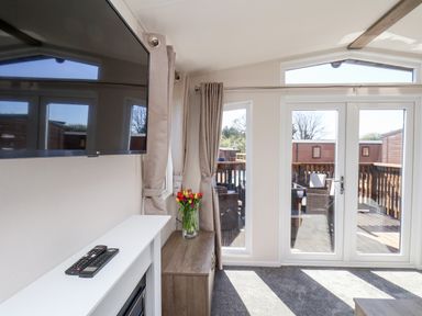 A living room with a wall-mounted TV a wooden cabinet with a vase of flowers and glass doors leading to an outdoor seating area at Mayflower Lodge in Runswick Bay near Staithes