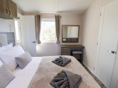 A bedroom with a bed with pillows and towels a window with curtains a door and a dressing table with a mirror at Mayflower Lodge in Runswick Bay near Staithes