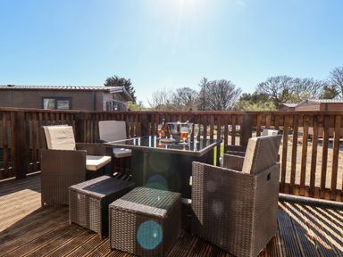 An outdoor deck with a glass table, four cushioned chairs, two footstools, and trees in the background at Mayflower Lodge in Runswick Bay near Staithes