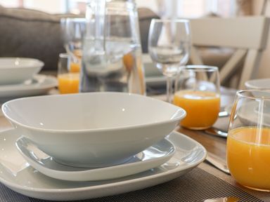 A dining table set with stacked white plates and bowls with glasses of orange juice and a water pitcher at Foxglove Lodge in Runswick Bay near Staithes