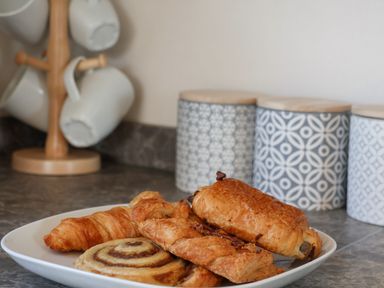 A plate with assorted pastries and a wooden mug tree with white mugs in a kitchen at Foxglove Lodge in Runswick Bay near Staithes