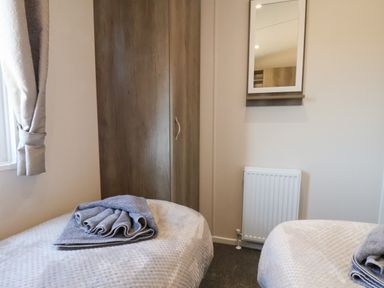 A bedroom with two beds, a wooden wardrobe, a radiator, a wall mirror, and a window with curtains at Foxglove Lodge in Runswick Bay near Staithes