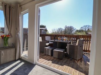 A wooden deck with outdoor wicker furniture including chairs and a table set seen through open glass doors from a carpeted room with curtains and a cabinet holding a vase of flowers at Foxglove Lodge Runswick Bay near Staithes
