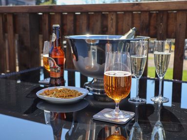 A glass of beer a bottle of beer a bowl of snacks a metal ice bucket with a bottle and two champagne glasses on a glass table on a wooden deck at Foxglove Lodge in Runswick Bay near Staithes