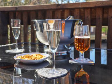 A glass of white wine an amber drink in a glass a bowl of snacks and a metal ice bucket on a glass table on a wooden deck at Tulip Lodge in Runswick Bay near Staithes