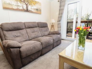 A living room with a brown sofa a wooden coffee table with flowers and a lamp near a window at Tulip Lodge in Runswick Bay near Staithes