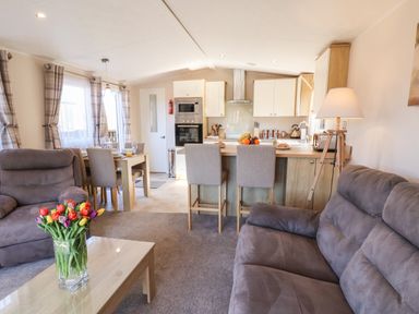 A living room with a coffee table with flowers two gray sofas a kitchen with stools and a dining table with chairs at Tulip Lodge in Runswick Bay near Staithes