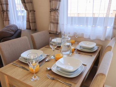 A dining table set for four with plates glasses of orange juice and a water jug in a room with beige curtains and upholstered chairs at Tulip Lodge in Runswick Bay near Staithes