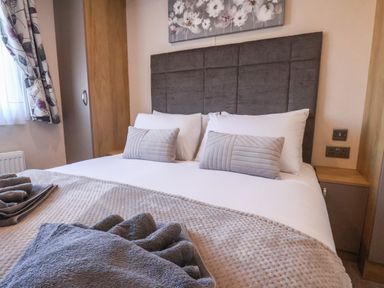 A bedroom with a bed made with white sheets gray pillows and towels next to a window with patterned curtains at Tulip Lodge in Runswick Bay near Staithes