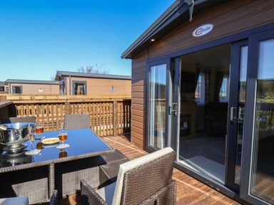 An outdoor patio with a glass table and wicker chairs next to a wooden building with sliding glass doors at Tulip Lodge in Runswick Bay near Staithes