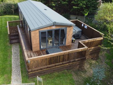 A wooden cabin with a gray metal roof and a fenced wooden deck in a grassy area at Tulip Lodge in Runswick Bay near Staithes