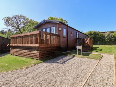 A wooden cabin with a deck and gravel driveway surrounded by grass and trees at Rose Lodge in Runswick Bay near Staithes