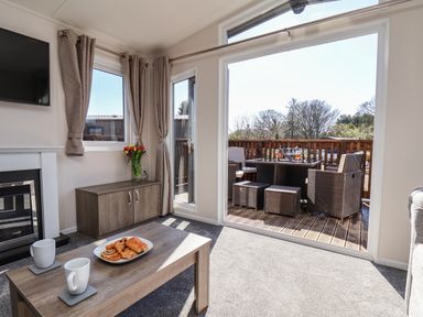 A living room with a coffee table holding pastries and mugs a vase with flowers on a cabinet and outdoor furniture visible through a large glass door at Honeysuckle Lodge in Runswick Bay near Staithes