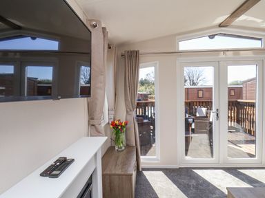 A living area with a wall mounted television remote controls flowers in a vase and glass doors leading to an outdoor seating area at Honeysuckle Lodge in Runswick Bay near Staithes